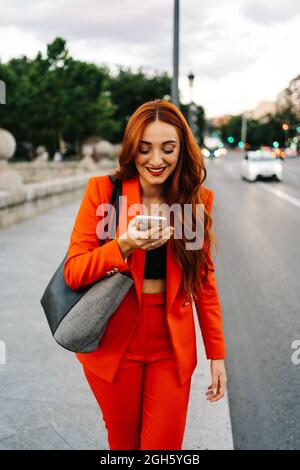 Lächelnde Frau mit roten Haaren und orangefarbenem Anzug, die auf dem Mobiltelefon eine Audionachricht aufzeichnet, während sie mit einem Freund in den sozialen Medien kommuniziert und hereinkommt Stockfoto