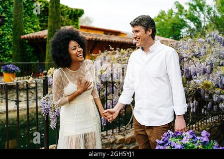 Inhalt multirassisches Paar, das die Hände hält, während es im Garten mit blühenden violetten Glyzinien-Blumen spazieren geht und das Wochenende gemeinsam genießt Stockfoto