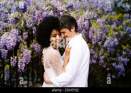 Seitenansicht eines liebevollen multirassischen Paares, das sich im Sommer im Park mit blühenden violetten Glyzinien-Blumen umarmt Stockfoto