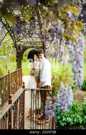 Seitenansicht eines sanften multirassischen Paares, das sich umarmt, während es mit geschlossenen Augen unter dem Bogen mit blühenden violetten Glyzinienblüten im Garten steht Stockfoto