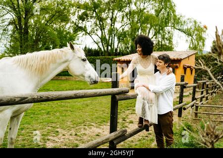 Mann umarmt schwarze Frau, die auf einem Holzzaun sitzt und im Sommer auf dem Land auf dem Weideland auf graues Pferd zugreift Stockfoto