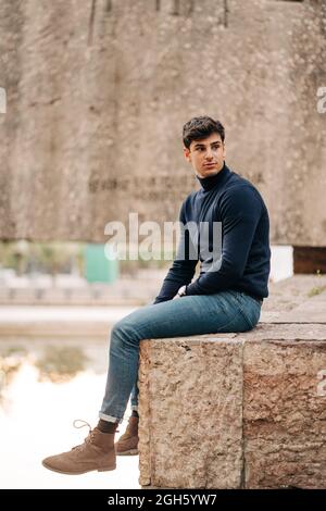 Seitenansicht eines jungen Mannes in trendigem Outfit, der auf einem Steinzaun in der Stadt sitzt und wegschaut Stockfoto