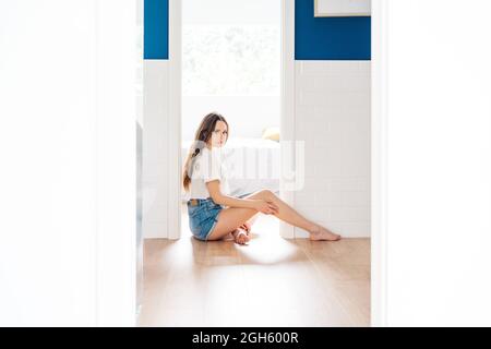 Seitenansicht einer jungen, stilvollen Frau in Denim-Shorts und weißem T-Shirt, die zu Hause am Boden in der Tür des Schlafzimmers sitzt und die Kamera anschaut Stockfoto