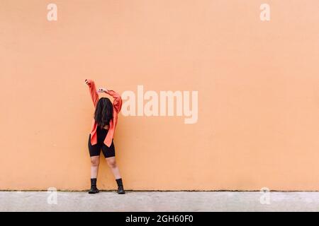 Ganzer Körper einer jungen anonymen Frau, die das Gesicht mit langen braunen Haaren bedeckt, die die Arme heben, während sie gegen eine orangefarbene Wand steht Stockfoto