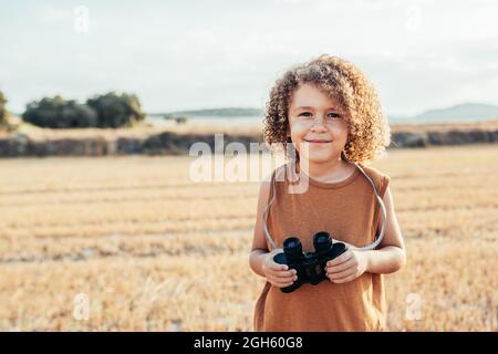 Entzücktes ethnisches Kind mit Afro-Frisur, das an einem sonnigen Tag im Sommer in getrockneten Fangen ein Fernglas hält und Spaß hat Stockfoto