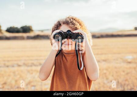 Entzücktes ethnisches Kind mit Afro-Frisur, das durch ein Fernglas schaute und den Sieg mit erhobenen Armen feierte, während es im Sommer in getrockneter Feichte stand Stockfoto