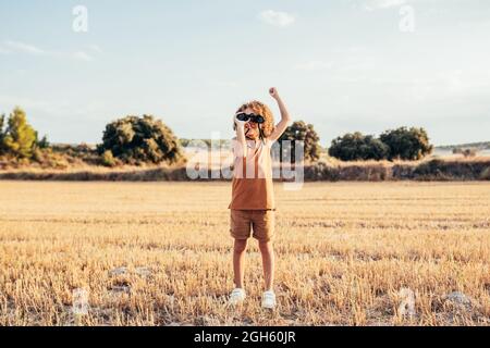 Entzücktes ethnisches Kind mit Afro-Frisur, das durch ein Fernglas schaute und den Sieg mit erhobenen Armen feierte, während es im Sommer in getrockneter Feichte stand Stockfoto