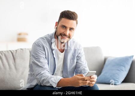 Ein gutaussehender Tausendjähriger Mann in Ohrstöpseln, der Smartphone benutzt und mit der Kamera lächelt, zu Hause auf dem Sofa sitzt und Platz kopiert Stockfoto