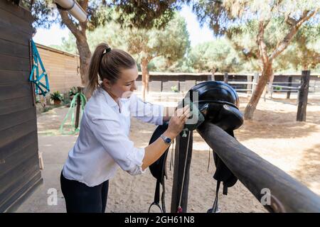 Seitenansicht der weiblichen Jockey waschen schwarzen Sattel während der Vorbereitung auf Reiten auf dem Bauernhof im Sommer Stockfoto