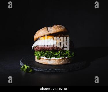 Leckerer Burger mit Ei auf Patty und frischer Salat auf Schiefertafel auf schwarzem Hintergrund im Studio serviert Stockfoto
