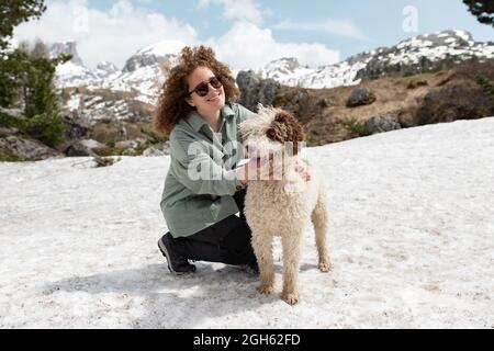 Frau spielt mit spanischem Wasserhund auf verschneiten Rasen in den Dolomiten, während sie an sonnigen Tagen im Winter Spaß hat Stockfoto