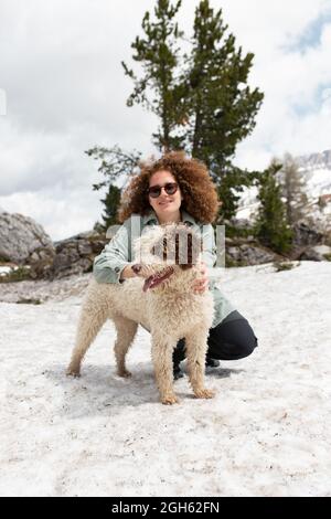 Frau spielt mit spanischem Wasserhund auf verschneiten Rasen in den Dolomiten, während sie an sonnigen Tagen im Winter Spaß hat Stockfoto