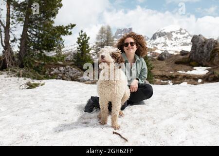 Frau spielt mit spanischem Wasserhund auf verschneiten Rasen in den Dolomiten, während sie an sonnigen Tagen im Winter Spaß hat Stockfoto