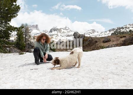 Frau spielt mit spanischem Wasserhund auf verschneiten Rasen in den Dolomiten, während sie an sonnigen Tagen im Winter Spaß hat Stockfoto