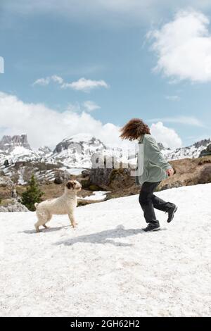 Seitenansicht einer nicht erkennbaren Hündin, die mit dem spanischen Wasserhund auf einer verschneiten Wiese in den Dolomiten spielt, während sie sich an einem sonnigen Wintertag amüsieren kann Stockfoto