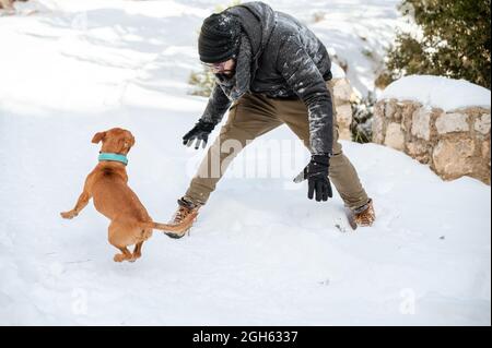 Männlicher Besitzer in warmer Kleidung, der im Winter mit einem niedlichen Hund im verschneiten Park spielt Stockfoto