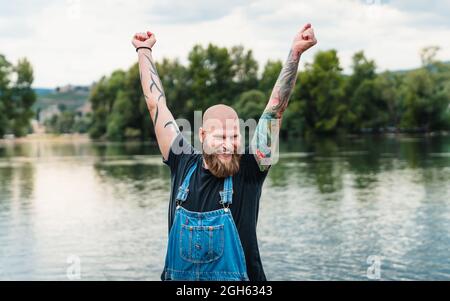 Glücklicher kahler Mann mit Bart und Tattoos in legerer Kleidung, der im Sommer mit erhobenen Armen in der Nähe des Sees und von grünen Bäumen steht Stockfoto