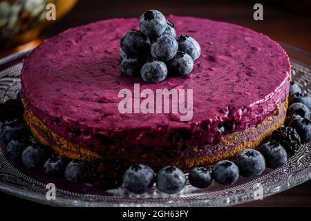 Köstliche Heidelbeer-Mousse-Torte mit violetter Creme, dekoriert mit frischen Beeren, serviert auf einem Glasständer auf einem dunklen Tisch Stockfoto