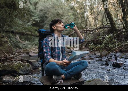 Durstiger männlicher Wanderer, der frisches Wasser aus der Flasche trinkt, während er auf einem Felsen in der Nähe des Flusses im Dschungel sitzt Stockfoto