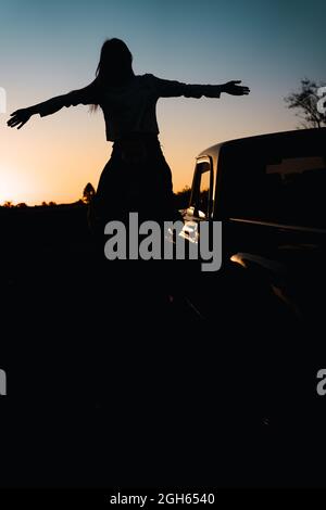Rückansicht der Silhouette einer nicht erkennbaren Frau mit ausgestreckten Armen, die auf den Schultern eines Mannes sitzen, der in der Nähe des Autos in der Natur auf dem Hintergrund des Sonnentauses steht Stockfoto