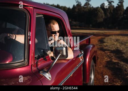 Selbstbewusster männlicher Fahrer mit Sonnenbrille sitzt im roten Retro-Pickup-Auto, das im Sommer auf einer Sandstraße auf dem Land geparkt ist Stockfoto