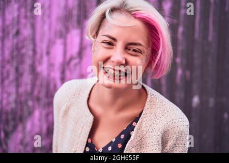 Charmante alternative weibliche mit gefärbten Haaren und in trendigen Kleidern stehen gegen violette Wand in der Stadt und Blick auf die Kamera Stockfoto