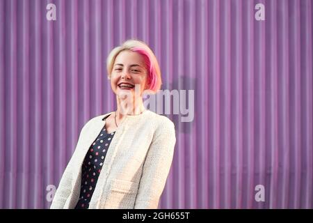 Charmante alternative weibliche mit gefärbten Haaren und in trendigen Kleidern stehen gegen violette Wand in der Stadt und Blick auf die Kamera Stockfoto