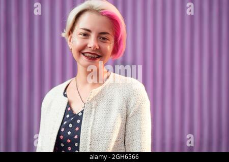 Charmante alternative weibliche mit gefärbten Haaren und in trendigen Kleidern stehen gegen violette Wand in der Stadt und Blick auf die Kamera Stockfoto