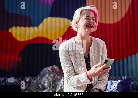 Seitenansicht einer trendigen alternativen Frau mit gefärbtem Haar, die auf einer Bank im Stadtgebiet der Stadt sitzt und in den sozialen Medien auf dem Handy surft Stockfoto