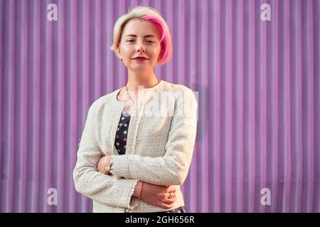 Charmante alternative weibliche mit gefärbten Haaren und in trendigen Kleidern stehen gegen violette Wand in der Stadt und Blick auf die Kamera Stockfoto