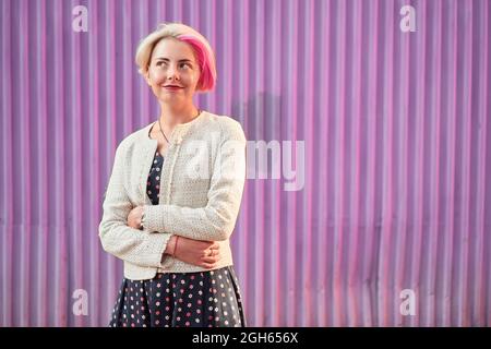 Charmante alternative weibliche mit gefärbten Haaren und in trendigen Kleidern stehen gegen violette Wand in der Stadt und wegschauen Stockfoto