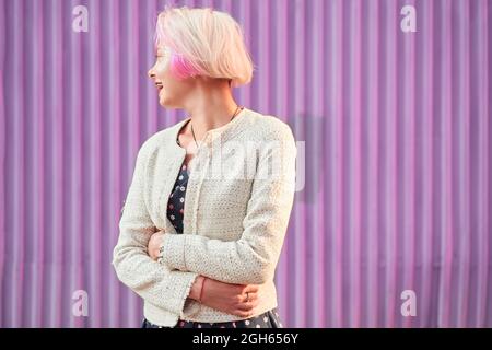 Charmante alternative weibliche mit gefärbten Haaren und in trendigen Kleidern stehen gegen violette Wand in der Stadt und wegschauen Stockfoto