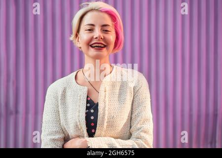 Charmante alternative weibliche mit gefärbten Haaren und in trendigen Kleidern stehen gegen violette Wand in der Stadt und Blick auf die Kamera Stockfoto