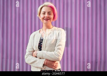 Charmante alternative weibliche mit gefärbten Haaren und in trendigen Kleidern stehen gegen violette Wand in der Stadt und Blick auf die Kamera Stockfoto