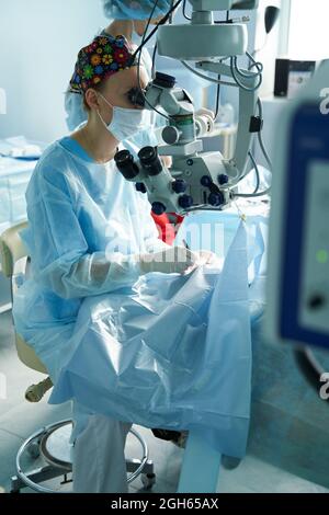 Aufmerksame Ärztin in einer chirurgischen Uniform und sterilen Maske, die durch das Mikroskop schaut, während sie das Auge eines nicht erkennbaren Patienten im Krankenhaus operiert Stockfoto