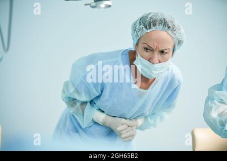 Konzentrierte reife Ärztin in steriler Uniform, die sich mit zusammengeklemmten Händen nach vorne lehnt und im Operationssaal wegschaut Stockfoto