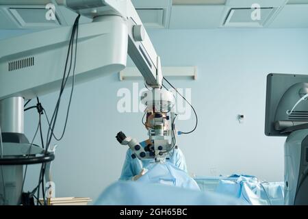 Aufmerksame Ärztin in einer chirurgischen Uniform und sterilen Maske, die durch das Mikroskop schaut, während sie das Auge eines nicht erkennbaren Patienten im Krankenhaus operiert Stockfoto