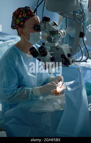 Aufmerksame Ärztin in einer chirurgischen Uniform und sterilen Maske, die durch das Mikroskop schaut, während sie das Auge eines nicht erkennbaren Patienten im Krankenhaus operiert Stockfoto