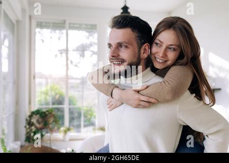 Lächelnde Frau, die zu Hause gegen die Fenster auf dem Huckepack auf ihrem Freund reitet Stockfoto