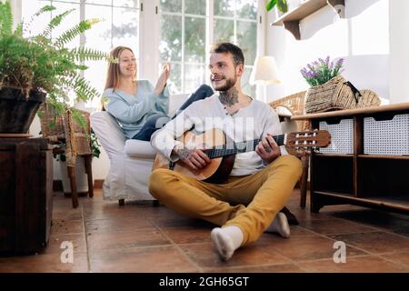 Tätowierte männliche Musikerin mit gekreuzten Beinen, die akustische Gitarre spielt, während die fröhliche weibliche Geliebte zu Hause im Sessel applaudiert Stockfoto