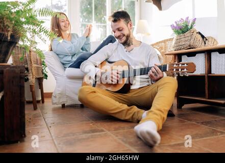 Tätowierte männliche Musikerin mit gekreuzten Beinen, die akustische Gitarre spielt, während die fröhliche weibliche Geliebte zu Hause im Sessel applaudiert Stockfoto
