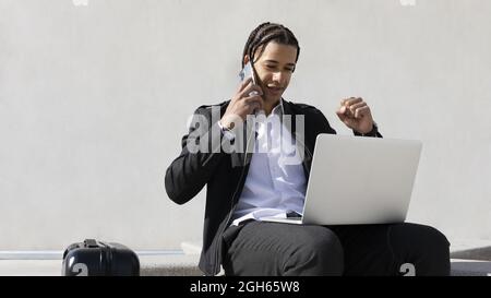 Positiver junger, gut gekleideter, ethnischer Unternehmer mit Zöpfen, die auf den Laptop-Bildschirm schauen und auf dem Mobiltelefon sprechen, während er über Geschäftsthema spricht Stockfoto