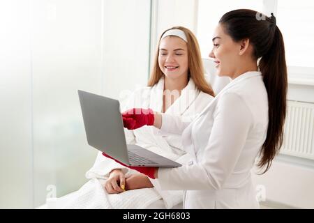 Ärztin, die mit dem Patienten arbeitet, während sie in der Klinik einen Laptop benutzt. Schönheitsarzt konsultiert den Patienten. Kosmetologe und Schönheitschirurgen Konzept Stockfoto