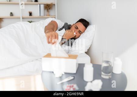 Kalt-, Rauch-, Coronavirus-Konzept. Kranker Kerl, der Servietten aus der Taschentuchbox nahm und niesen laufende Nase, die im Bett lag Stockfoto