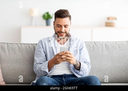 Junger Mann in Ohrhörern, der auf dem Smartphone chattet und zu Hause auf dem Sofa sitzt, freier Platz. Technologie und Gadgets Konzept Stockfoto