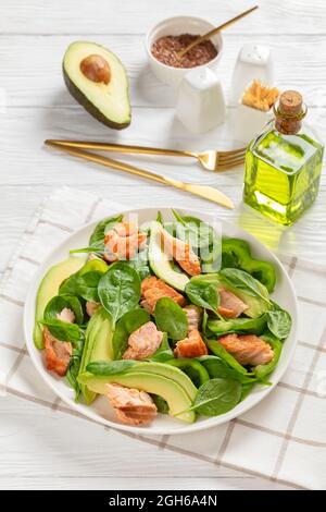 Lachs Spinat Avocado und grüner Pfeffer Salat auf einem weißen Teller mit goldener Gabel und Messer auf einem weißen Holztisch, mediterrane gesunde Ernährung, vertikal Stockfoto