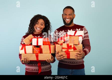 Glückliches afroamerikanisches Paar mit vielen Weihnachtsgeschenken und Weihnachtspullover auf blauem Hintergrund Stockfoto