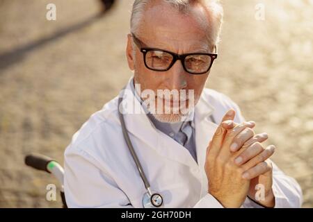 Nahaufnahme eines nachdenklichen, behinderten älteren Arztes im Rollstuhl, der an einem sonnigen Tag im Freien einen Labormantel und eine Brille trägt Stockfoto