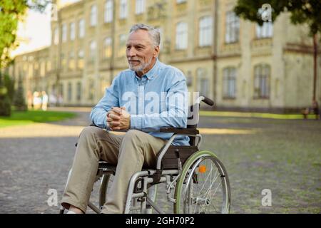 Nachdenklicher, reifer Mann, der sich im Rollstuhl erholt, schaut weg, während er Zeit allein im Park in der Nähe des Krankenhauses verbringt Stockfoto