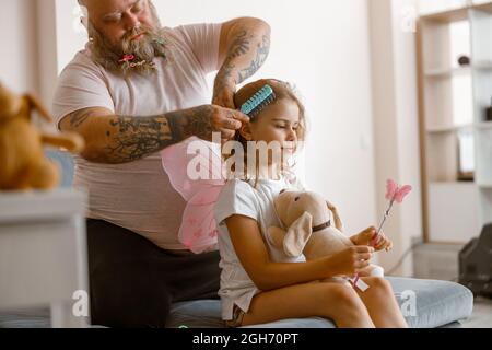 Vaty mit Scrunchies im Bart bürstet Haare von niedlichen kleinen Tochter zu Hause Stockfoto